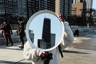 Person holding a camera in one hand and a mirror with a highrise building inside in the other. They’re wearing grey jeans, a white fluffy jacket, and white Nike sneakers.