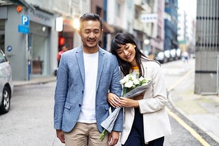 A photo of a happy Asian couple walking down the street. The woman is holding a bouquet of white flowers.