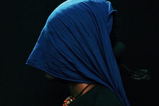 a person in profile view against a black background. They are wearing a vibrant blue headscarf or veil that completely covers their head and face. The person is dressed in a dark green top or sweater and has a colorful beaded necklace visible at their neck, featuring orange, red, and multi-colored beads. The composition is striking in its contrast between the deep blue of the head covering and the darkness of the background, creating a contemplative, mysterious atmosphere.