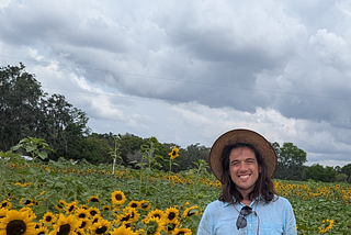 Ron Rivers standing in a sunflower farm and wearing a sun hat, sky blue colour half sleeves shirt and sea green shirts with a wide smile on his face looking at you.