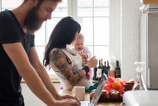 Young mother holding her newborn baby in the kitchen while the father works on his laptop in the foreground.