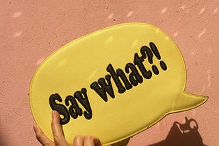A hand holding up a yellow handbag in the form of a speech bubble, with the text: “Say what?!”