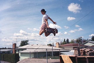 A teenage boy in a t-shirt, shorts, and a baseball cap holds his skateboard to his feet after launching himself in the air from a plywood ramp, framed against the sky above the homes of a working-class neighborhood in 1980s Spokane.