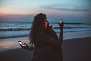 A couple hugs at the beach during sunset while holding their phones out and looking at them.