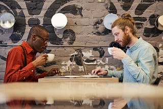 A black man and a white man in front of their laptops sipping coffee directly across from each other at a cafe table.