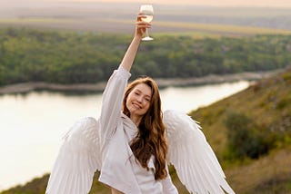 Angel with wavy brunnette hair and white clothing raising a glass of white wine. She is standing on a hill overlooking a river. The background is blurred, but the lighting gives a sense of serence peace to the overall picture.