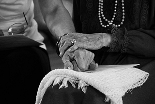 A black and white photograph of an elderly person resting her hand on a younger person’s arm in a gesture of loving support.