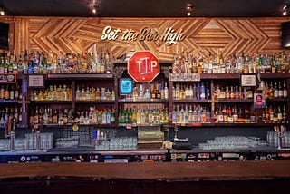 A bar stocked w/ liquor. Gold letters near the ceiling say “Set the bar high”; below is a stop sign spraypainted to say “Tip”