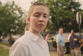 A close-up shot of a woman. We can see from her shoulders up. She is wearing a plain white shirt. Her body is aimed to the right, but her face is turned towards to the camera. She has blonde hair. Behind her, blurred out, are two FBI officers, and behind them a car. The woman is not smiling, her face is solemn and expressionless.