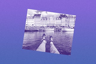 Filtered image of a person’s legs dangling over water with European buildings in the background.