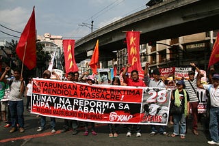 A crowd of protesters holding vertical red flags and carrying a large banner with Tagalog writing.