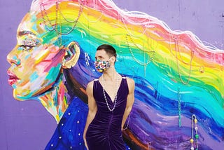 A person wearing a jeweled face mask posing in a sleeveless dark-purple dress in front of a rainbow mural of Laverne Cox.