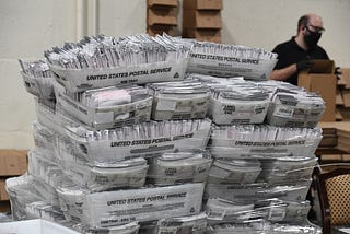 Stacks of mail cartons full of ballots being sorted by a person in the background wearing a face mask.