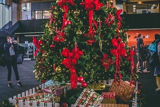 Large Christmas tree in a public space. Tree is decorated in large red bows. People appear to be shopping nearby and one man is taking pictures of the tree.