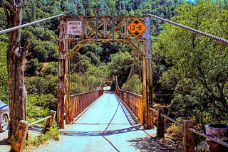 Old rusty single-lane gravel-covered suspension bridge with weight limit limit sign reading maximum of three-tons and an orange warning sign noting maximum height of thirteen feet, eight inches. The image was taken straight on down the center of the roadway with the thick bradded wires projecting out from the bridge to out of sight past the photographer. The bridge road empties onto a single road into a steep hillside thickly covered by trees and brush. The true length distance is deceiving.