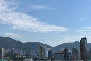 A blue sky with swirling, fluffy clouds over a view of Hong Kong with buildings in front of mountains.
