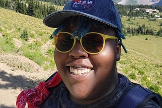 Author on top of a mountain with green hills rolling behind them leading toward a mountain top crested with clouds on a sunny day. Aigner Loren Wilson is smiling wearing yellow sunglasses and a jean NASA hat. Author’s life. Life of a publishing speculative fiction author. Speculative Fiction author. Aigner Loren Wilson