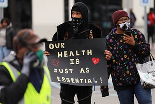 A photo of Amazon workers protesting. One is holding up a sign that says “Our health is just as essential.”