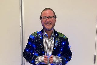 The author has a giant grin on his face. He’s wearing glasses, has a beard, and his hair is pulled back into a bun. He’s wearing a black suit with blue LED lights, flowers, leaves, and beads sewn in. Underneath is a shiny white shirt covered in little squares.
