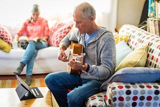 An older man learning guitar by watching an online tutorial on his tablet.