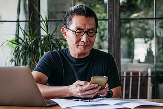 An older man texts on his cell phone while sitting down at a table with paperwork and his laptop.