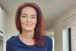 This is a headshot of the author. She is Caucasian, thin and has red hair swept to the right. She has blue/green eyes and is wearing a navy blue long sleeve fitted shirt. In the background is a white kitchen with a windowed door on the right.