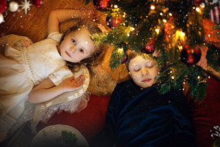 Two young children, seen from above, lie on a parquet floor beneath a decorated Christmas tree with warm lights. One gazes into the camera, the other has their eyes closed. Sparkly snowfall overlays the scene.