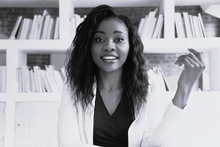 Photo of woman in white lab coat sitting at a desk. She has a look of surprise on her face, as if she can’t believe she has to explain what she is explaining.