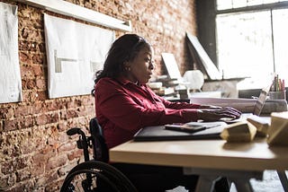 A Black businesswoman in a wheelchair works at her desk.