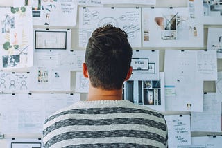 A person looking at a wall covered with paper sheets of paper containing sketches and annotated pictures.