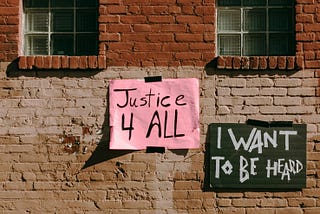 Two handmade protest signs taped to a weathered brick wall below green-shuttered windows. The left sign on pink paper reads ‘Justice 4 All’ in black lettering, while the right sign on dark green paper reads ‘I Want To Be Heard’ in white text.