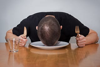 A person holding a fork and knife at a dinner table, resting their head on an empty plate.