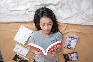 Girl laying on her bed reading with books scattered around her
