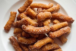 A photo of a plate piled high with homemade golden fried churros, tube-shaped dough that’s dusted in sugar and cinnamon