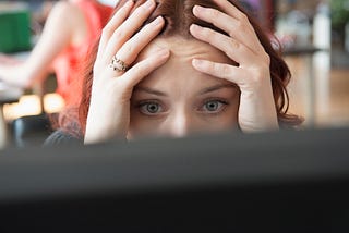 A photo of a stressed woman with her hands on the sides of her face, looking at her computer screen.