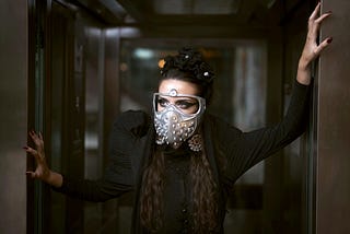 A woman wearing a dark costume and a mask is posing inside an elevator.