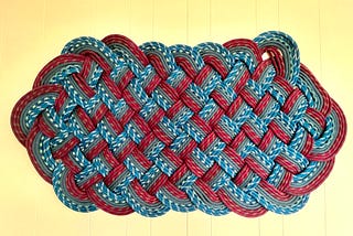 Color photo of a cream wooden wall background on which hangs a large ‘knotted’ sailing artwork in pale blue and dark red paracord rope, rectangularly wider than it is tall.