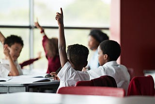 A photo of a young black boy with his hand raised at his desk.