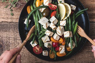 Hands using wooden utensils to toss a bowl of green beans, cherry tomatoes, and grilled tofu cubes in sauce/vinaigrette.