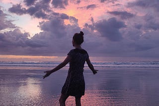 A woman in a dress standing ankle-deep in the ocean with arms spread in front of a pink, purple, and blue sunset.