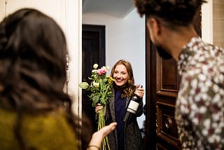 A woman holding a bouquet and a wine bottle in their neighbor’s doorway.