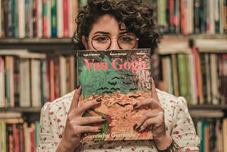 A woman is standing in front of shelves of books, holding a “Van Gogh” book. She is hiding her mouth behind the book, with her eyes and glasses peeking out over the top.
