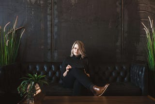 A woman wearing black is sitting on a black couch while holding a tablet.