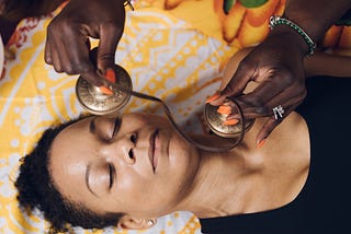 A holistic practitioner holding bells over the face and neck of a person lying on their back with their eyes closed.