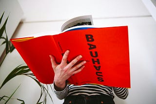 Photograph of someone in a striped artist’s jersey reading a big, bold red hardback book about the Bauhaus