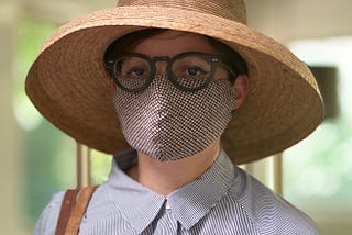 A person wearing a button-up shirt, round glasses, a wide-brimmed straw hat, and a face mask with a diamond pattern.