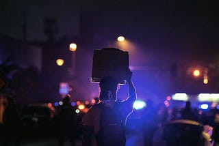 A dark nighttime photo of a protestor holding up a sign with police lights in the background.
