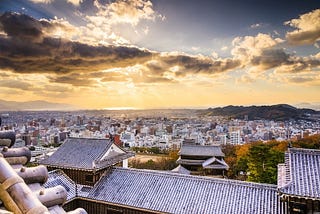 Sunset view over Matsuyama City from the rooftops of Matsuyama Castle, with golden light spilling across the skyline and the Seto Inland Sea glowing in the distance.