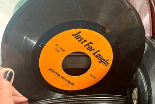 A stack of black 45rpm records, with the female author’s left thumb with matching black nail polish holding up the top record showing the orange background title “Just For Laughs”.