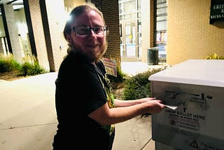 The author is putting his ballot in a drop box. He is wearing a black t-shirt and has dark-rimmed glasses. It’s nighttime. He looks anxious and excited.